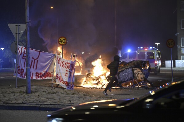 A person runs past a burning car and a banner reading, 