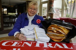 In this Thursday, Oct. 31, 2019, photo, Sue Dillon poses in her home in Carmel, Ind., with some of the petitions gathered to change Indiana time zone. Dillon became a campaigner for changes to the state's time choice after a teenager was fatally struck in 2009 while running to catch a school bus in the early morning darkness near her home. (AP Photo/Michael Conroy)