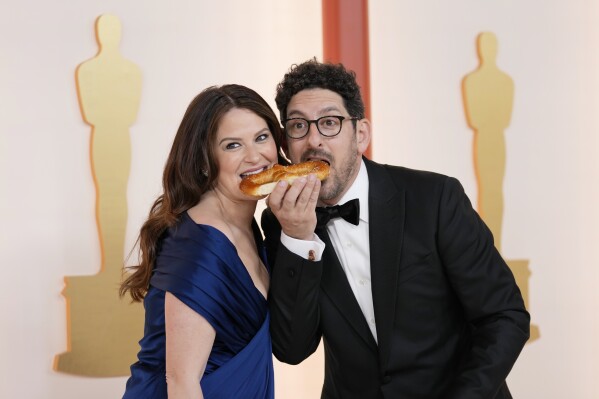 Katie Lowes, left, and Adam Shapiro eat pretzels on the carpet at the Oscars on Sunday, March 12, 2023, at the Dolby Theatre in Los Angeles. (AP Photo/Ashley Landis)
