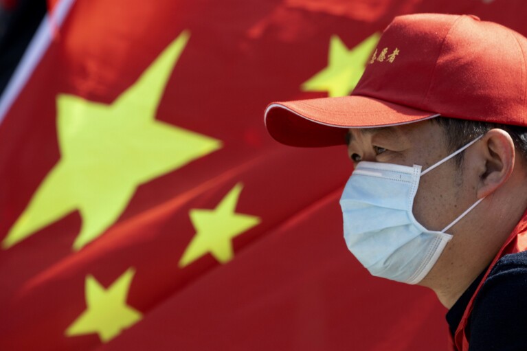 FILE - A volunteer looks out near a Chinese national flag during a farewell ceremony for the last group of medical workers who came from outside Wuhan to help the city during the coronavirus outbreak in Wuhan in central China's Hubei province on April 15, 2020. The hunt for COVID-19 origins has gone dark in China. An AP investigation drawing on thousands of pages of undisclosed emails and documents and dozens of interviews found feuding officials and fear of blame ended meaningful Chinese and international efforts to trace the virus almost as soon as they began, despite years of public statements to the contrary. (AP Photo/Ng Han Guan, File)