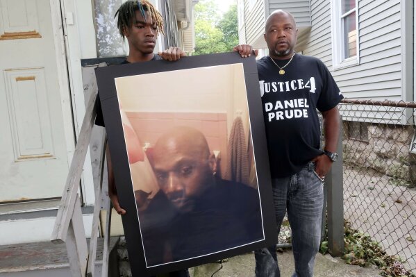 FILE — In this Sept. 3, 2020 file photo, Joe Prude, brother of Daniel Prude, right, and his son Armin, stand with a picture of Daniel Prude in Rochester, N.Y. Newly released transcripts show that a grand jury investigating the police suffocation death of Daniel Prude last year in Rochester, New York, voted 15-5 to clear the three officers involved in his restraint of a criminally negligent homicide charge sought by prosecutors. (AP Photo/Ted Shaffrey, File)