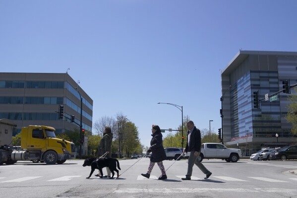 FILE - Maureen Reid, left, and her guide dog, Gaston, cross the intersection of Wood Street and Roosevelt Avenue with Sandy Murillo, center, and Geovanni Bahena, relying on an audible signal for the blind, on April 26, 2023, in Chicago. The U.S. Census Bureau wants to change how it asks people about disabilities, and some advocates are complaining that they were not consulted enough on what amounts to a major overhaul in how disabilities would be defined by the federal government. (AP Photo/Charles Rex Arbogast, file)