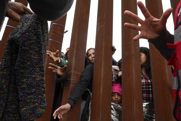 Migrants reach through a border wall for clothing handed out by volunteers in San Diego, as they wait to apply for asylum for entry into the United States on May 12, 2023. (AP Photo/Gregory Bull)