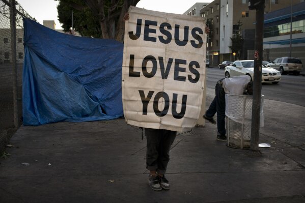 Samuel Raymond, 50, holds up a banner saying "Jesus Loves You" on a sidewalk as a passing homeless man reaches into a trash can in search of anything of value Saturday, Nov. 4, 2017, in the Skid Row area of downtown Los Angeles. "The Lord is my shelter," said Raymond, who has been homeless for nearly 20 years. (AP Photo/Jae C. Hong)