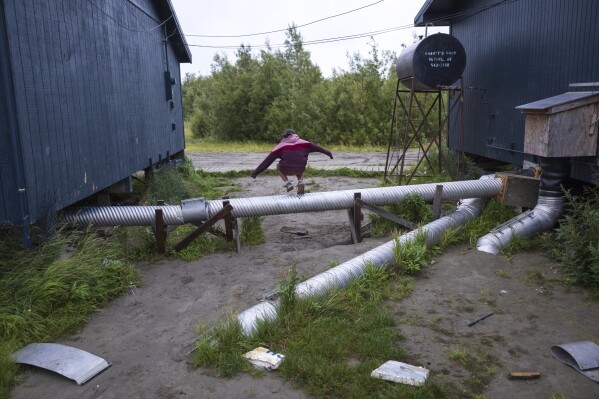 Andi Nicholai leaps over a pipe between village administrative buildings, Sunday, Aug. 20, 2023, in Akiachak, Alaska. (AP Photo/Tom Brenner)