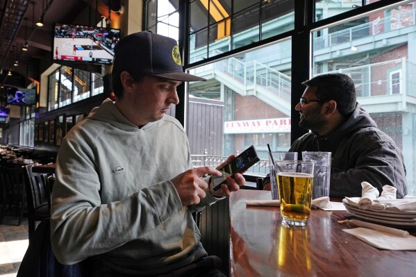 FILE - Taylor Foehl, left, of Boston, looks at the mobile betting app on his phone after placing a wager, while watching a men's college basketball game at the Cask 'N Flagon sports bar, Friday, March 10, 2023, near Fenway Park in Boston. A company that most of the legal U.S. sports betting industry uses to verify that its customers are where they say they are reported on Wednesday, Sept. 13, a record number of transactions over the first weekend of the NFL season. (AP Photo/Charles Krupa, File)