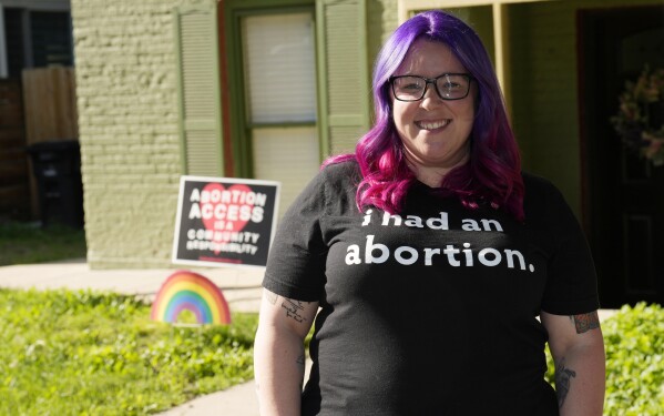 Leah Dean, a native of the Texas Panhandle, poses outside her home Monday, July 3, 2023, in Denver. Americans are segregating by their politics at a rapid clip, helping fuel the greatest divide between the states in modern history. In Texas, Dean had been scared to fly an abortion rights banner outside her house. Now in Denver, an LGBTQ+ pride flag flies above the banner in front of her house that proclaims, “Abortion access is a community responsibility.” (AP Photo/David Zalubowski)