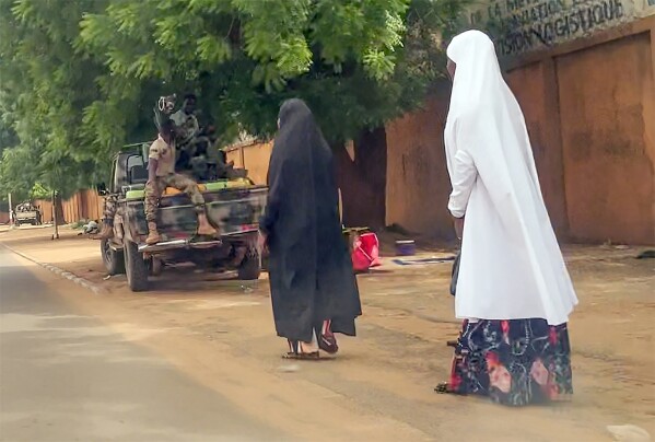 In this image made from video, two women walk past soldiers in Niamey, Niger, Friday, July 28, 2023. The general who led a coup in Niger defended the takeover on state television and asked for support from the nation and international partners, as concerns grew that the political crisis could set back the country's fight against jihadists and increase Russia's influence in West Africa. (AP Photo)