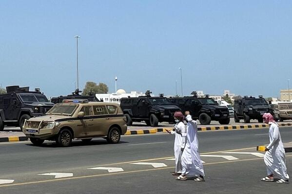 People walk near riot police cars in Sohar, Oman, Tuesday, May 25, 2021. On Tuesday, dozens of protesters angry over firings and the poor economy of Oman marched in Sohar, a city some 200 kilometers northwest of the capital, marking a third day of demonstrations in the typically subdued sultanate. (AP Photo)