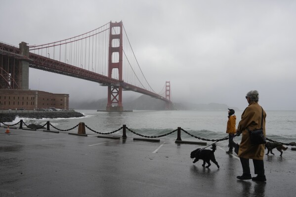People walk their dogs during a break between rain showers near the Golden Gate Bridge and Fort Point in San Francisco, Tuesday, Dec. 19, 2023. (AP Photo/Eric Risberg)