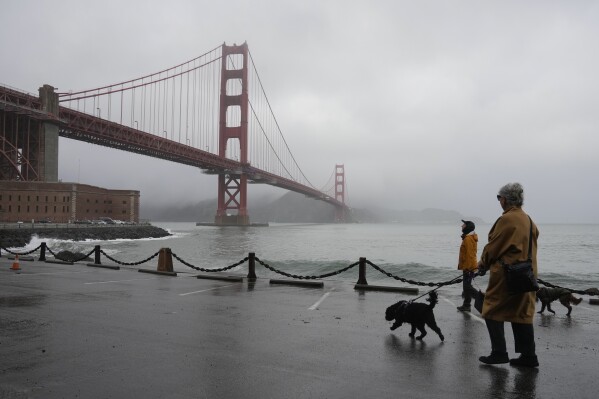 People walk their dogs during a break between rain showers near the Golden Gate Bridge and Fort Point in San Francisco, Tuesday, Dec. 19, 2023. (AP Photo/Eric Risberg)