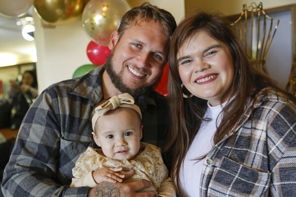 Alan Shurtleff, left, Morgan Shurtleff, right, and their daughter, 1-year-old Cora Dibert, pose for a photo at The Bridge Church, Saturday, Dec. 2, 2023, in Mustang, Okla. When Cora went for a routine blood test in October, the toddler brought along her favorite new snack: a squeeze pouch of WanaBana cinnamon-flavored apple puree. Within a week, the family got an alarming call. The test showed that the 1-year-old had lead poisoning, with nearly four times as much lead as the level that raises concern. (AP Photo/Nate Billings)
