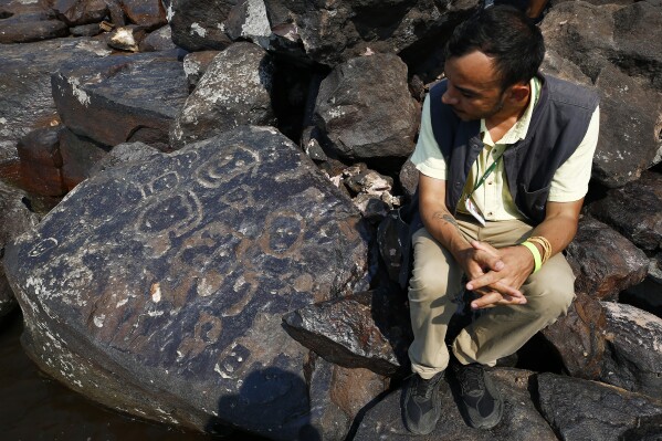 Archaeologist Jaime Oliveira sits next to rock paintings at the Ponta das Lajes archaeological site, in the rural area of Manaus, Brazil, Saturday, Oct. 28, 2023. The archaeological site was exposed following a drought in the Negro River, unveiling rock paintings that, according to archaeologists, date back between 1,000 and 2,000 years. (AP Photo/Edmar Barros)