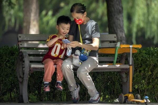FILE - A woman wearing a face mask and a child look at a cellphone as they sit on a bench at a public park in Beijing, on June 2, 2022. As the week-long Lunar New Year holidays in China draw near with promises of feasts and red envelopes stuffed with cash, children have yet another thing to look forward to - one extra hour of online games each day. (AP Photo/Mark Schiefelbein, File)