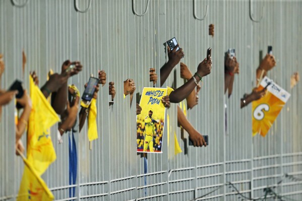 Fans carry a poster of Chennai Super Kings' MS Dhoni and take pictures through a fence as they wait for the Indian Premier League cricket match between Chennai Super Kings and Punjab Kings to begin in Chennai, India, Wednesday, May 1, 2024. (AP Photo/R. Parthibhan)