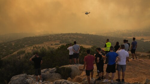 A firefighting helicopter flies through smoke as people look on in Mandra west of Athens, on Tuesday, July 18, 2023. In Greece, where a second heatwave is expected to hit Thursday, three large wildfires burned outside Athens for a second day. Thousands of people evacuated from coastal areas south of the capital returned to their homes Tuesday when a fire finally receded after they spent the night on beaches, hotels and public facilities. (AP Photo/Petros Giannakouris)