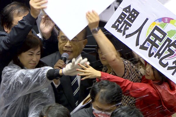 In this image made from video, Taiwanese Premier Su Tseng-chang holds a microphone as opposition party lawmakers from the Nationalist party (KMT) block his attempt to speak during a parliament session in Taipei, Taiwan, Friday, Nov. 27, 2020. Taiwan's lawmakers got into a fist fight and threw pig guts at each other Friday over a soon-to-be enacted policy that would allow imports of U.S. pork and beef. The banner at right reads: “Democratic Progressive Party feeds people with ractopamine pork.” (FTV via AP)