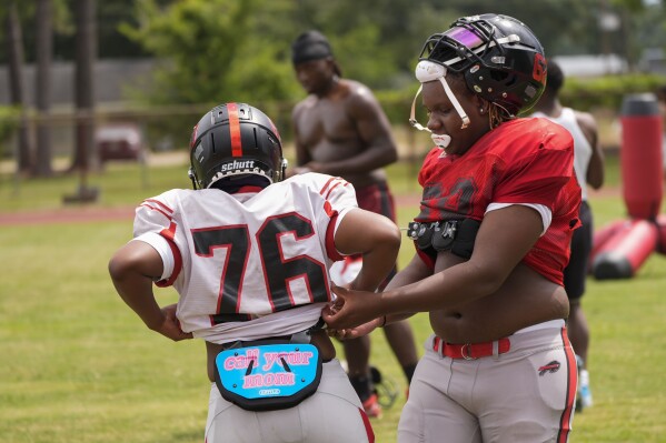 Keiland Burns, left, wears a protective pad that reads "call your mom" as teammate Jamie Allen, right, helps him with his gear at the start of football practice at Baker High in Baker, La., Wednesday, Aug. 28, 2024. (AP Photo/Gerald Herbert)