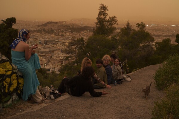 Tourists play with a cat at Lycabettus hill as the city of Athens with the ancient Acropolis hill is seen at the background, on Tuesday, April 23, 2024. The Acropolis and other Athens landmarks took on Martian hues Tuesday as stifling dust clouds blown across the Mediterranean Sea from North Africa engulfed the Greek capital. (AP Photo/Petros Giannakouris)
