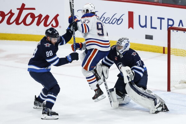 Edmonton Oilers' Connor McDavid (97) jumps in front of Winnipeg Jets goaltender Connor Hellebuyck (37) as Nate Schmidt (88) defends during the third period of an NHL hockey game Thursday, Nov 30, 2023, in Winnipeg, Manitoba. (John Woods/The Canadian Press via AP)