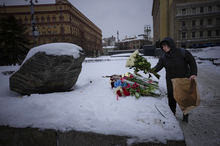 Moteris padeda gėles Aleksejaus Navalno garbei prie paminklo, didelės Soloveckio salų uolos, kur buvo įkurta pirmoji Gulago politinių kalėjimų sistemos stovykla su istoriniu Federalinės saugumo tarnybos (FSB, sovietinio KGB įpėdinio) pastatu. fone, Maskvoje, Rusijoje, šeštadienio rytą, 2024 m. vasario 17 d. (AP nuotr./Aleksandras Zemlianichenko)