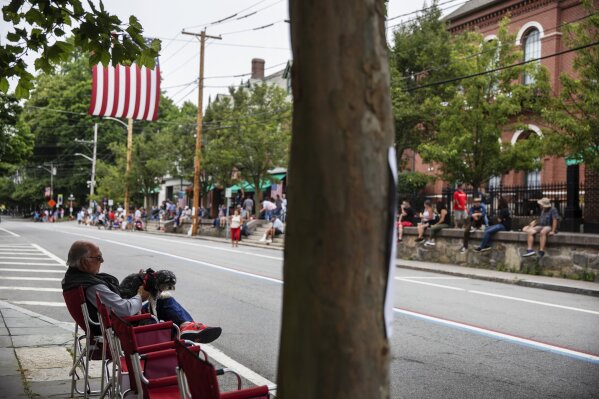 Mike Howard sits with his dog Molly while waiting for the Fourth of July parade to pass his home Saturday, July 4, 2020, in Bristol, R.I. Howard said he normally places chairs out on the sidewalk at 3:30 a.m. to hold a spot but didn't have to this year with far fewer people attending. The town, which lays claim to the nation's oldest Independence Day celebration in the country, held a vehicle-only scaled down version of its annual parade Saturday due to the coronavirus pandemic. (AP Photo/David Goldman)