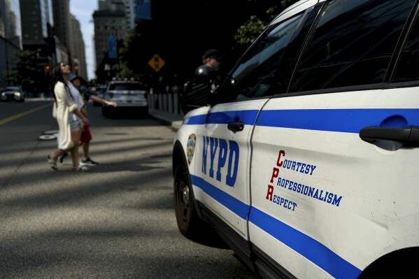 Pedestrians pass in front of a New York Police Department patrol car as they cross Greenwich Street, Wednesday, July 10, 2024, in New York. The New York Police Department will stop promoting “Courtesy, Professionalism, Respect” on the exterior of its patrol cars, replacing the long-standing three-word motto with a new slogan that reads: “Fighting crime, protecting the public.” (AP Photo/Julie Jacobson)