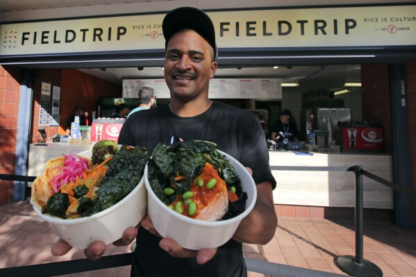 FILE - This Aug. 30, 2019 photo shows chef JJ Johnson posing with two signature rice bowls, one salmon and one vegetable, outside his Field Trip counter-service restaurant kiosk on the food court at the US Open tennis championships in New York. Johnson's new cookbook 