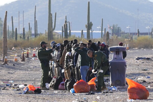Members of the U.S. Border Patrol and U.S. Customs and Border Protection organize a group of migrants as hundreds of migrants gather along the border Tuesday, Dec. 5, 2023, in Lukeville, Ariz. The U.S. Border Patrol says it is overwhelmed by a shift in human smuggling routes, with hundreds of migrants from faraway countries like Senegal, Bangladesh and China being dropped in the remote desert area in Arizona. (AP Photo/Ross D. Franklin)