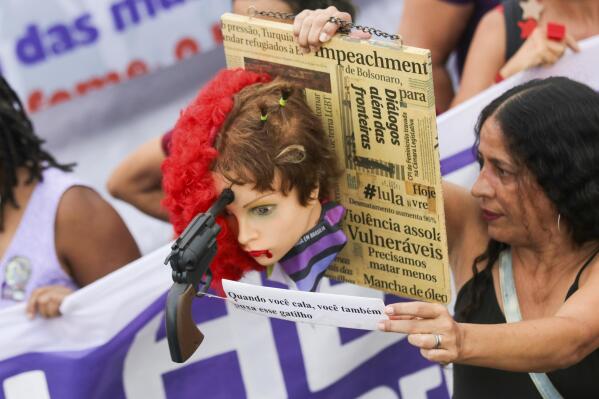 A woman holds the head of a mannequin with a gun pointed at its head and a sign that reads "When you shut up, you also pull that trigger" during a a march marking International Women's Day, in Brasilia, Brazil, Wednesday, March 8, 2022. (AP Photo/Gustavo Moreno)