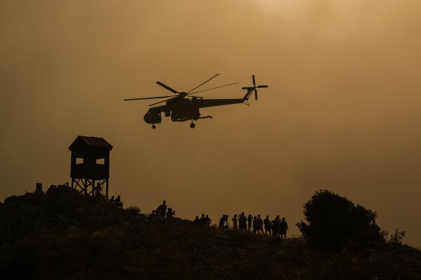 A firefighting helicopter flies through smoke as people watch, at the fire in Mandra west of Athens, on Tuesday, July 18, 2023. In Greece, where a second heatwave is expected to hit Thursday, three large wildfires burned outside Athens for a second day. Thousands of people evacuated from coastal areas south of the capital returned to their homes Tuesday when a fire finally receded after they spent the night on beaches, hotels and public facilities. (AP Photo/Petros Giannakouris)