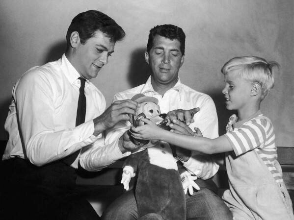 Jay North, young star of the new "Dennis the Menace" series, right, attempts to handcuff Dean Martin, center, as they play with a stuffed toy with Tony Curtis on Sept. 15, 1959. (AP Photo, File)