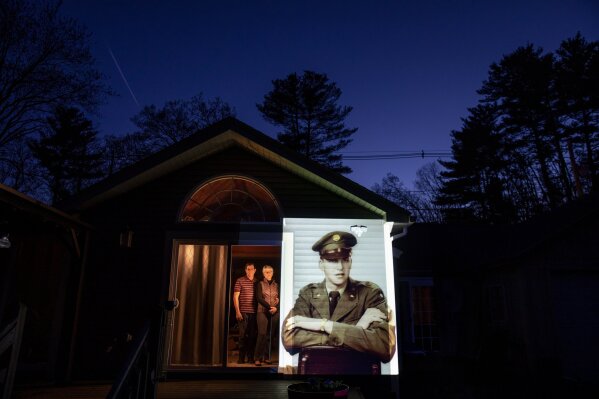 An image of veteran Roy Benson is projected onto the home of his daughter, Robin Benson Wilson, left, as she looks out a doorway with her husband, Donald, in Holland, Mass., Wednesday, May 13, 2020. Benson, a U.S. Army veteran and resident of the Soldier's Home in Holyoke, Mass., died from the COVID-19 virus at the age of 88. Seeking to capture moments of private mourning at a time of global isolation, the photographer used a projector to cast large images of veterans on to the homes as their loved ones are struggling to honor them during a lockdown that has sidelined many funeral traditions. (AP Photo/David Goldman)