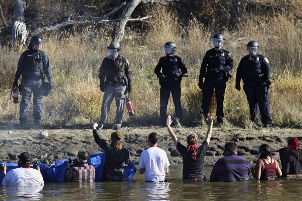 FILE - Protestors demonstrating against the expansion of the Dakota Access Pipeline wade in cold creek waters confronting local police, near Cannon Ball, N.D., Nov. 2, 2016. A court fight over whether the federal government should cover North Dakota's $38 million in costs of responding to the lengthy protests of the Dakota Access oil pipeline years ago near its controversial river crossing will continue as a judge said the case is “ripe and ready for trial.” The trial is set to begin Feb. 15 in Bismarck. (AP Photo/John L. Mone, File)