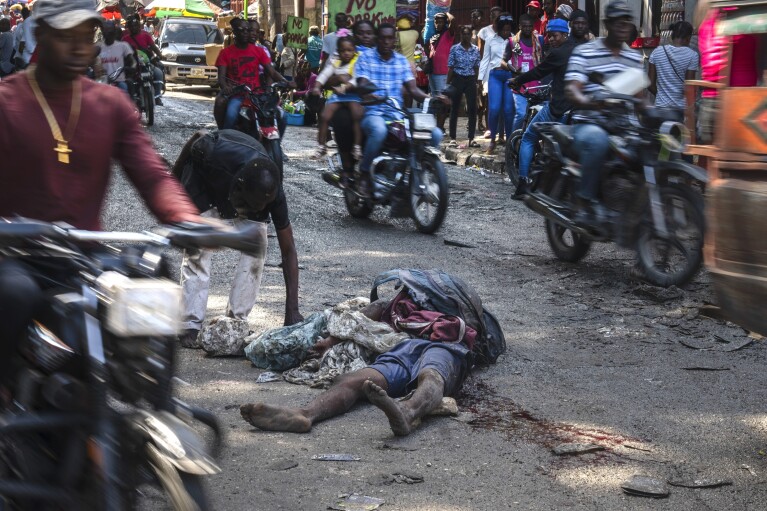 ARCHIVO - Un cuerpo yace en la calle mientras los viajeros, incluida una niña en la parte trasera de una motocicleta, arriba en el centro, se abren paso por el barrio de Petion-Ville de Puerto Príncipe, Haití, el 22 de abril de 2024. Cuando era joven Los haitianos están cada vez más expuestos a la violencia y el país está realizando un esfuerzo más amplio para disipar un tabú de larga data sobre buscar terapia y hablar sobre salud mental. (Foto AP/Ramón Espinosa, Archivo)