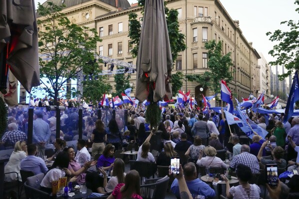 People sit on a cafe terrace while others wave flags during a rolling HDZ party rally in downtown Zagreb, Croatia, Sunday, April 14, 2024. Croatia this week holds a parliamentary election following a campaign that was marked by heated exchanges between the country's two top officials, creating a political crisis in the Balkan country, a European Union and NATO member state. (AP Photo/Darko Bandic)