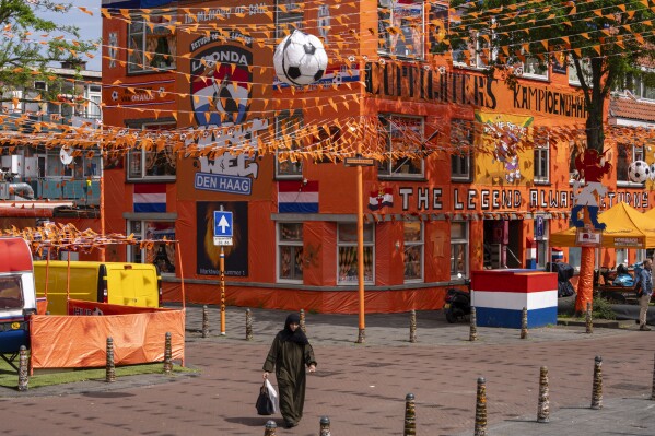 A women walks past inflatable soccer balls, orange tarp, orange bunting, and Dutch national flags as she walks along Marktweg street in The Hague, Netherlands, Thursday June 13, 2024, one day ahead of the start of the Euro 2024 Soccer Championship. The Marktweg is one of several streets in the Netherlands that get an all-encompassing orange facelift during European Championships and World Cups when the national team, known as Oranje after the Dutch royal family and the color of their shirts, are playing. (AP Photo/Peter Dejong)