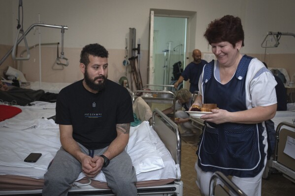 A nurse brings lunch for a wounded professional soldier from Medellín, Colombia, who goes by the call sign of Checho, 32, in a hospital in Ukraine on Wednesday, Dec. 20, 2023. Checho joined the Ukrainian armed forces to help fight Russia. After two years of war, Ukraine is looking for ways to replenish depleted ranks and the Colombian army volunteers are a welcome addition. (AP Photo/Efrem Lukatsky)