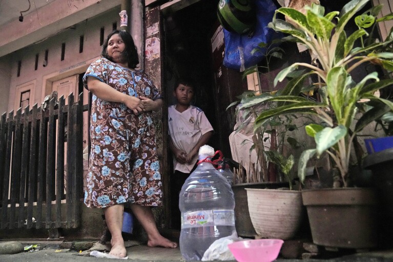 Devi Putri Eka Sari, a stay-at-home mother, stands outside her house with her son Satria Fareji, near bottles she uses to collect water, in Jakarta, Indonesia, Wednesday, March 13, 2024. Instead Sari, like millions of Indonesians across the country, buy water in large refillable containers or single-use packaged plastic bottles. (AP Photo/Dita Alangkara) (AP Photo/Dita Alangkara)