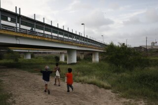FILE - In this June 26, 2019 file photo, migrant children walk with their families along the Rio Grande, as pedestrian commuters use the Puerta Mexico bridge to enter Brownsville, Texas, seen from Matamoros, Tamaulipas state, Mexico. The U.S. this week started having immigrant children held in Houston appear before a judge based in Atlanta, in what advocates say is a pilot that could portend a nationwide expansion of video hearings for kids. While the government would not confirm its plans, advocates warned of a greater burden being placed on detained immigrant children, many of whom are not yet teenagers and don’t have guaranteed access to an attorney.  (AP Photo/Rebecca Blackwell, File)