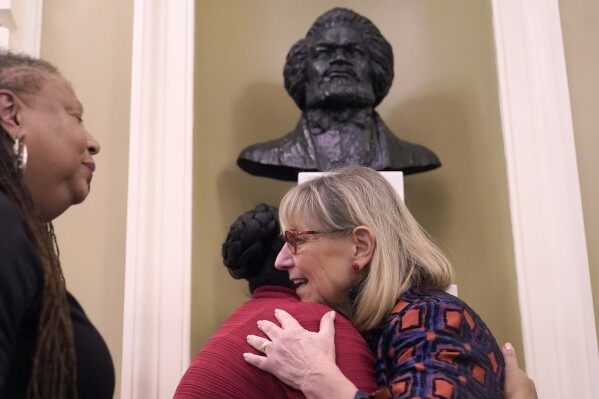 Karen Spilka, president of the Massachusetts Senate, front right, embraces Noelle Trent, President and CEO of the Museum of African American History, behind center, as L'Merchie Frazier, member of the State House Art Commission, left, looks on Wednesday, Feb. 14, 2024, during the unveiling of a permanent bust of famed abolitionist Frederick Douglass, top, in the Senate Chamber at the Statehouse, in Boston. It's the first bust of an African American to be permanently added to the Massachusetts Statehouse and the first bust to be added to the Senate Chamber in more than 125 years. (AP Photo/Steven Senne)