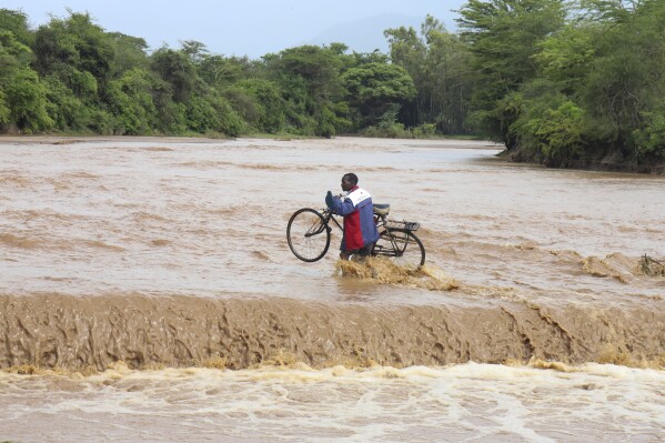 A man carrying his bicycle crosses through a flooded Muuoni River, where 8 people are said to have drowned overnight while crossing the river at Mukaa area, Makueni county, Kenya's Eastern region, Friday Nov. 24, 2023. Flood-related death toll is going higher everyday as heavy rains continue to pound across East Africa. (AP Photo)