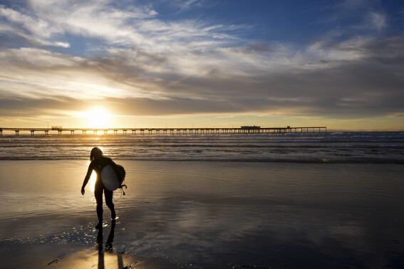 A surfer walks in front of the Ocean Beach pier as the sun sets, Tuesday, Jan. 30, 2024, in San Diego. Rising seas, frequent storms take toll on California's iconic piers, threatening beach landmarks. (AP Photo/Gregory Bull)