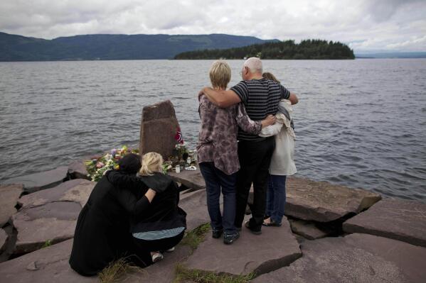 FILE - In this Monday, July 25, 2011 file photo, relatives of a victim gather to observe a minute's silence on a campsite jetty on the Norwegian mainland, across the water from Utoya island, seen in the background, where people have been placing floral tributes in memory of those killed in the shooting massacre on the island.  At 3.25 p.m. on July 22, 2021, a ray of sun should have illuminated the first of 77 bronze columns on a lick of land opposite Utoya island outside Oslo. Over the next 3 hours and 8 minutes, it would have brushed each column in turn, commemorating every person killed by right-wing terrorist Anders Breivik. But on the ten-year anniversary of the terror, the memorial remains a construction site.  (AP Photo/Matt Dunham, File)