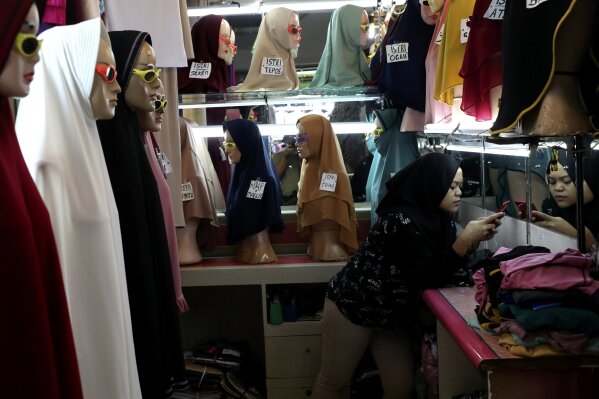 An employee uses her mobile phone as she waits for customers at a shop selling Muslim women's headscarves at a market in Jakarta, Indonesia, Wednesday, Nov. 11, 2020. As companies race to develop a COVID-19 vaccine and countries scramble to secure doses, questions about the use of pork products — banned by some religious groups — has raised concerns about the possibility of disrupted immunization campaigns. (AP Photo/Tatan Syuflana)