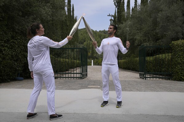 The first torchbearer, Greek Olympic gold medalist Stefanos Douskos, right, passes the flame to the first French torchbearer, three-time Olympic medalist Laure Manaudou, near the monument to Pierre de Coubertin, in the background, after the official ceremony ignition of the flame.  for the Paris Olympic Games, at the site of Ancient Olympia, Greece, Tuesday, April 16, 2024. (AP Photo/Thanassis Stavrakis)