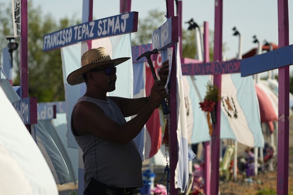 Christian Rodriguez tacks a poem to a cross that is part of a makeshift memorial at the site where officials found dozens of people dead in an abandoned semitrailer containing suspected migrants last summer, Tuesday, June 27, 2023, in San Antonio. U.S. authorities announced the arrests of four more people in connection to the smuggling deaths of 53 migrants, including eight children, who were left in a tractor trailer in the scorching Texas summer. (AP Photo/Eric Gay)