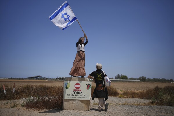 An activist stands on a concrete block signaling border ahead as she waves an Israeli flag towards trucks loaded with humanitarian aid for the Gaza Strip in southern Israel, Wednesday, May 21, 2025. (AP Photo/Ohad Zwigenberg)