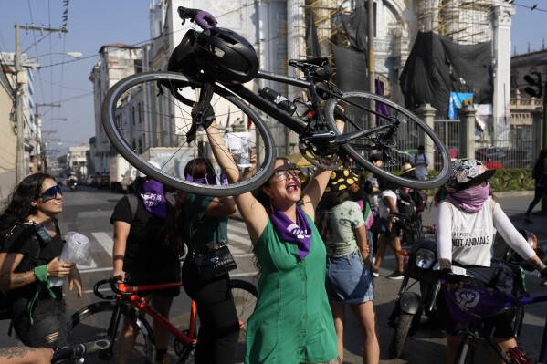 A woman lifts her bicycle in order to stop traffic during a march marking International Women's Day in Guatemala City, Friday, March 8, 2024. (AP Photo/Moises Castillo)