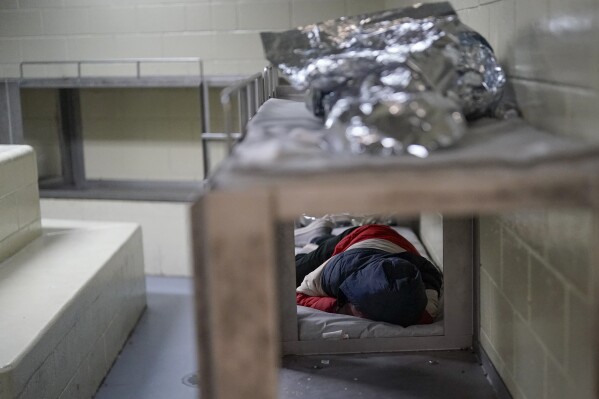 A person sleeps in a holding area Friday, Dec. 15, 2023, at a Border Patrol station in Ajo, Ariz. (AP Photo/Gregory Bull)